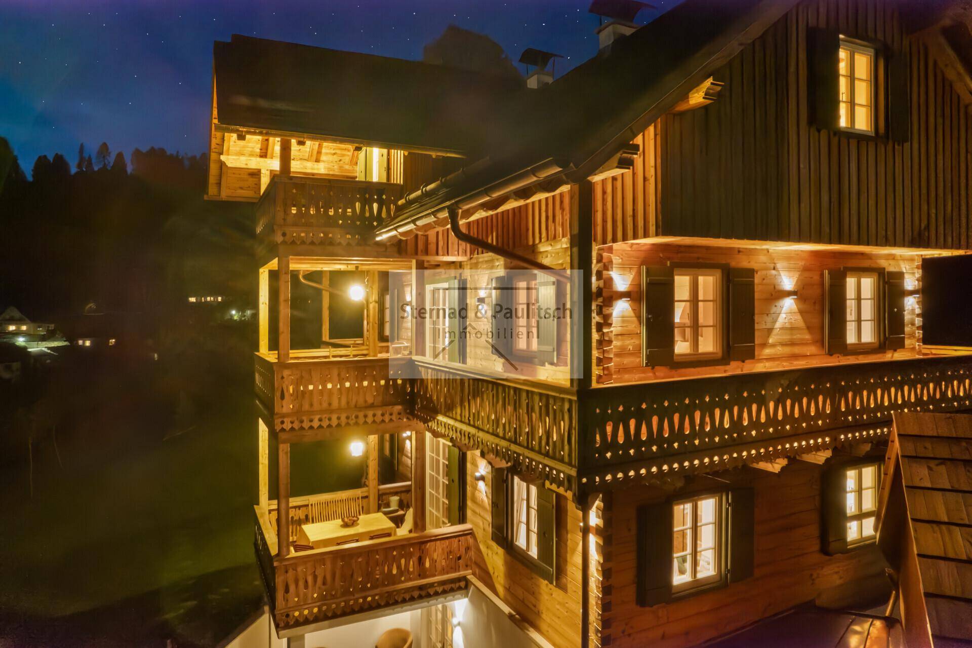Detailansicht des beleuchteten Holzchalets bei Nacht, mit sichtbaren Balkonen und Fenstern.