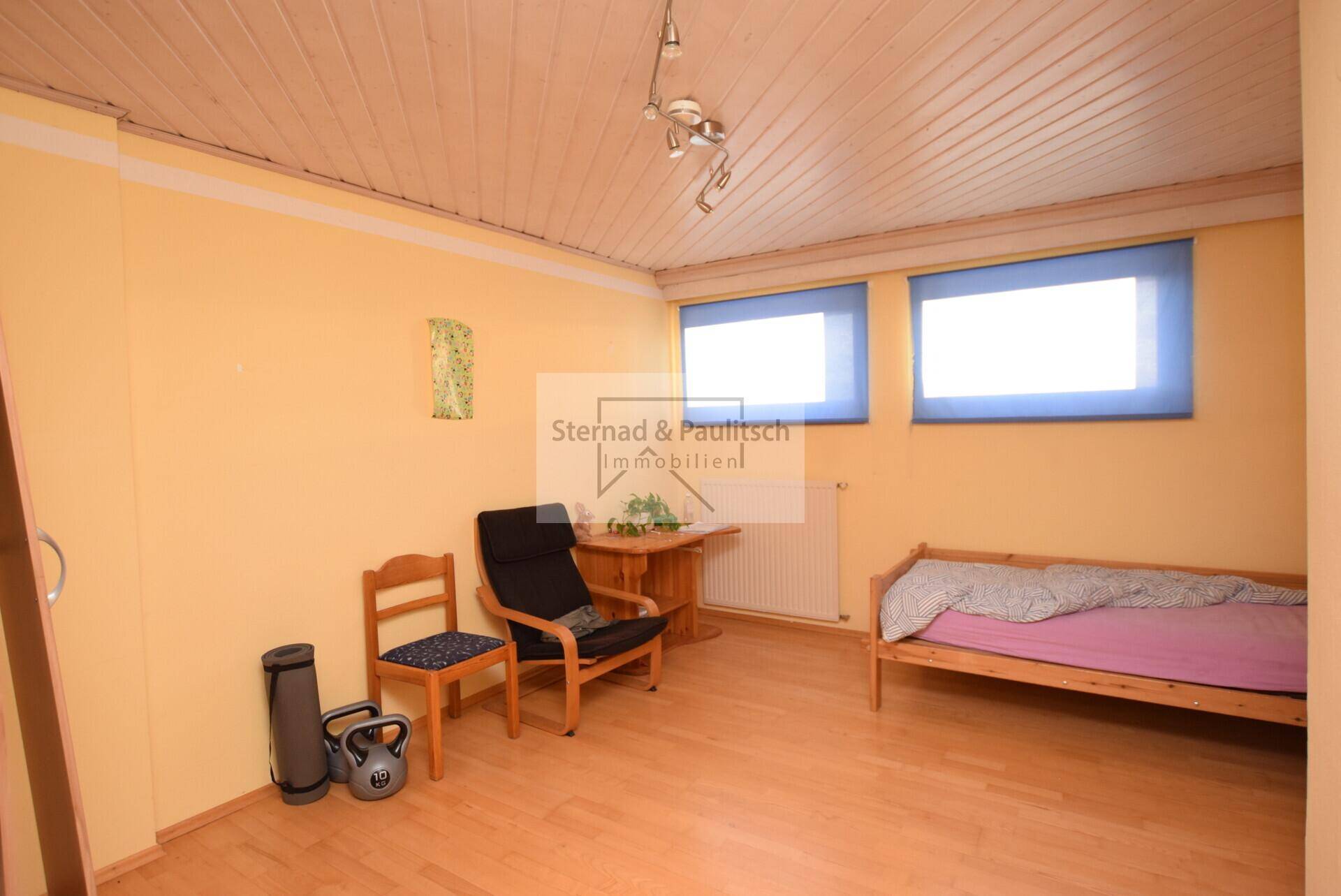 Helles Schlafzimmer mit gelben Wänden, Holzboden und zwei Fenstern, einfach eingerichtet.