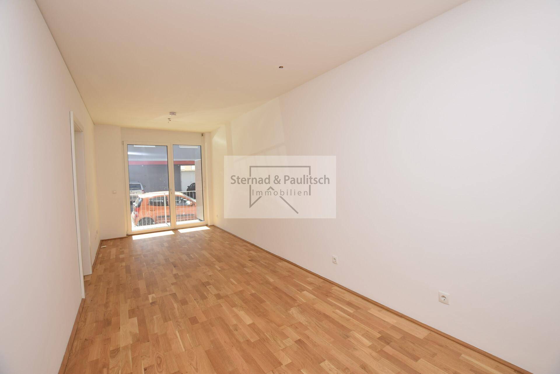 Leeres Zimmer mit Parkettboden und großem Fenster, bietet Zugang zu einem Außenbereich.