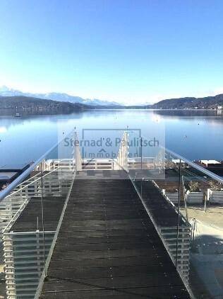 Moderner Holzsteg mit Glasgeländer, der sich über den See erstreckt und Bergblick bietet.