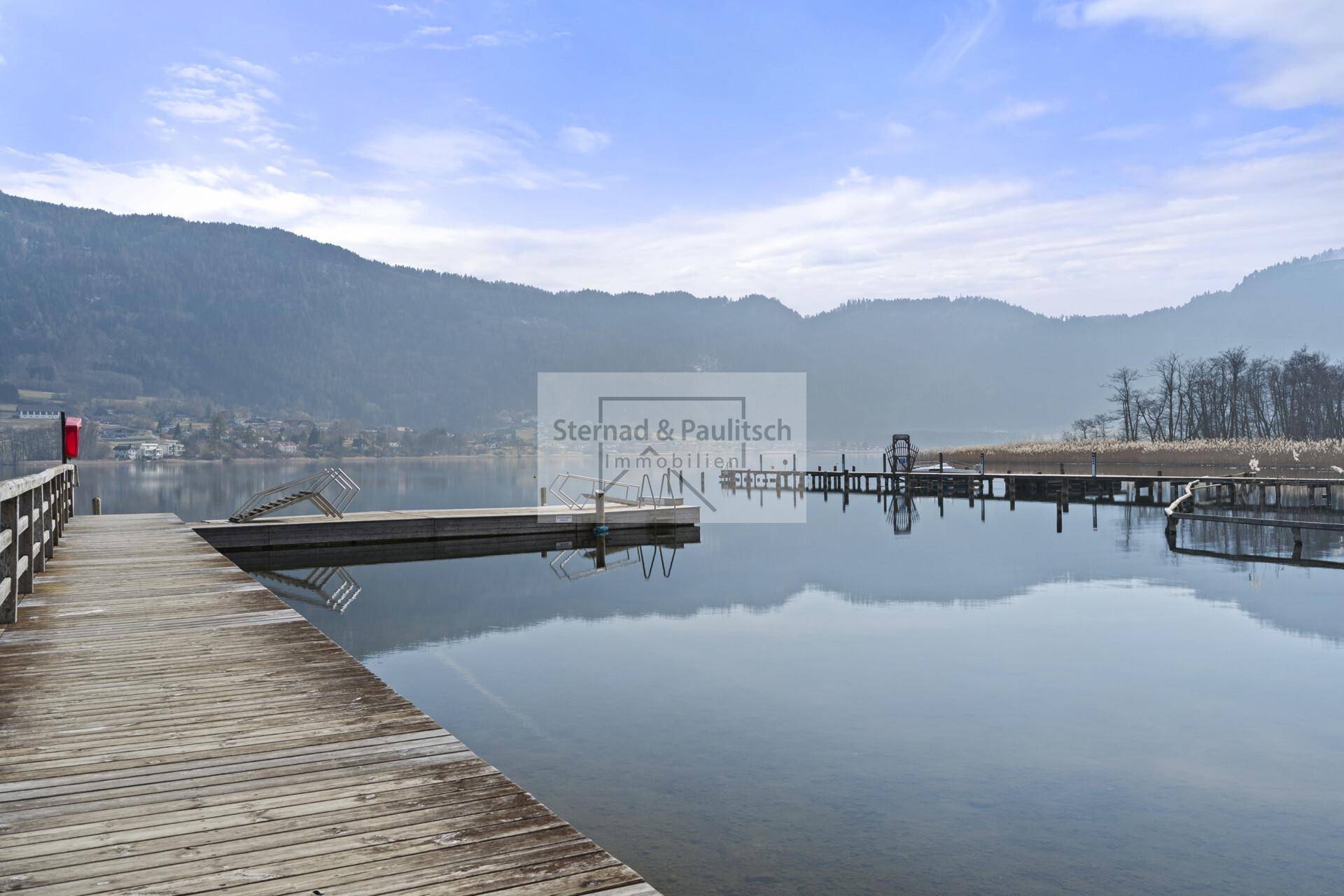 Ruhiger See mit Holzstegen und Bergen im Hintergrund unter einem weiten Himmel.