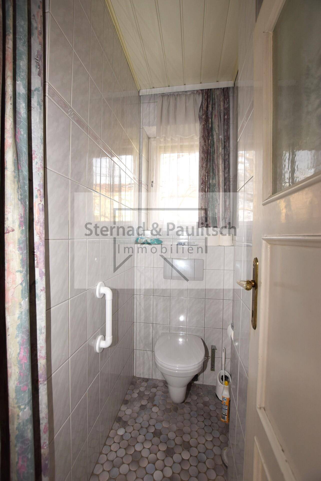 Kleines WC mit Fliesenboden, Toilette und Haltegriff.