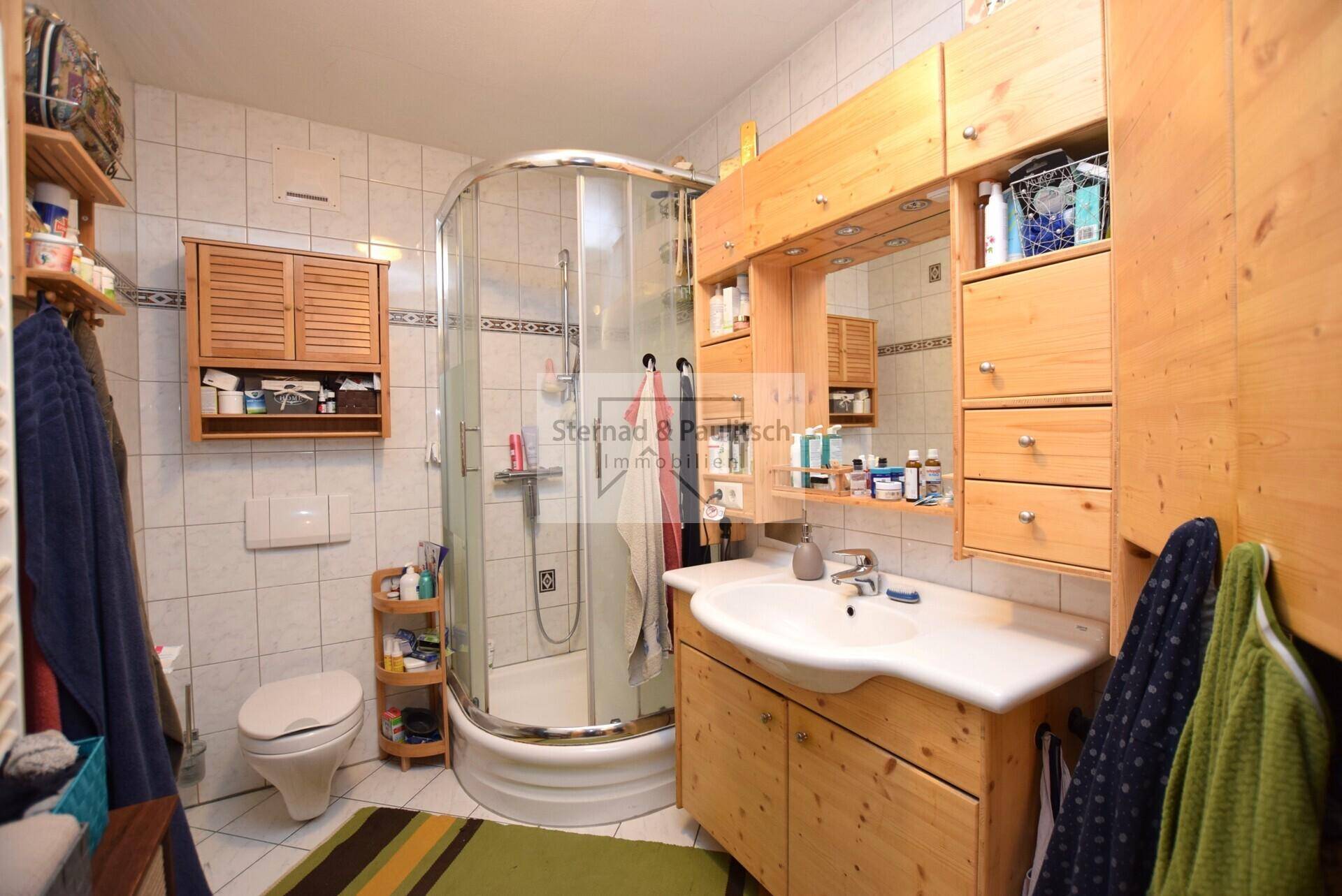 Badezimmer mit Duschkabine, Holzschränken, WC und hellen Fliesen.