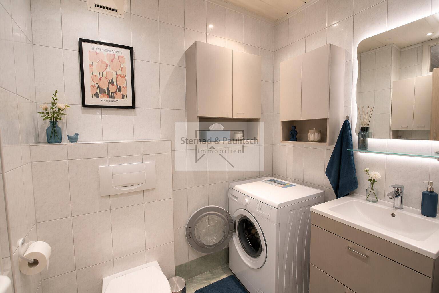 Modernisiertes Badezimmer mit WC, neuem Waschbecken und beleuchtetem Spiegel.