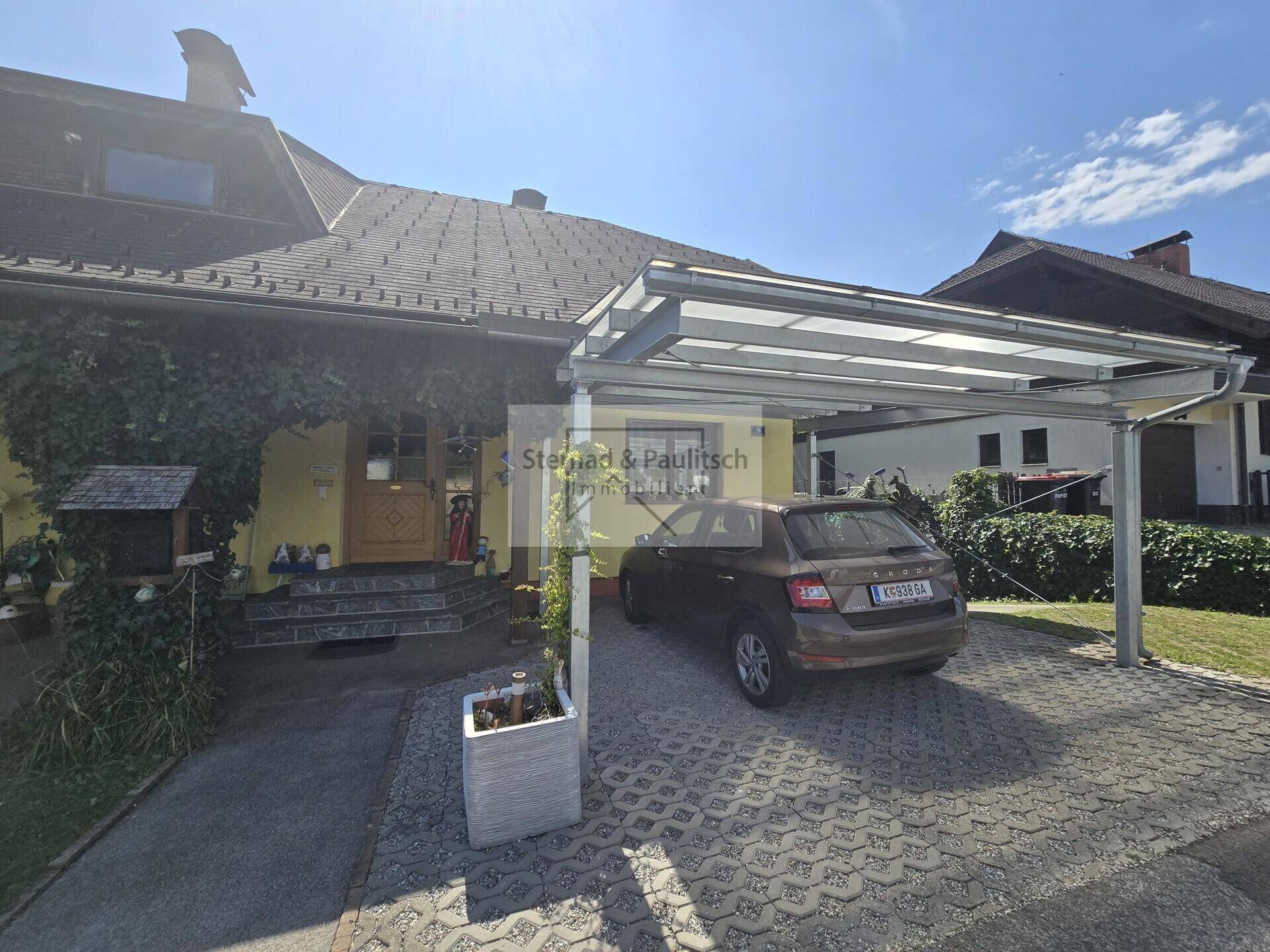 Hausfassade mit überdachtem Carport und gepflasterter Einfahrt, umgeben von Grünflächen.