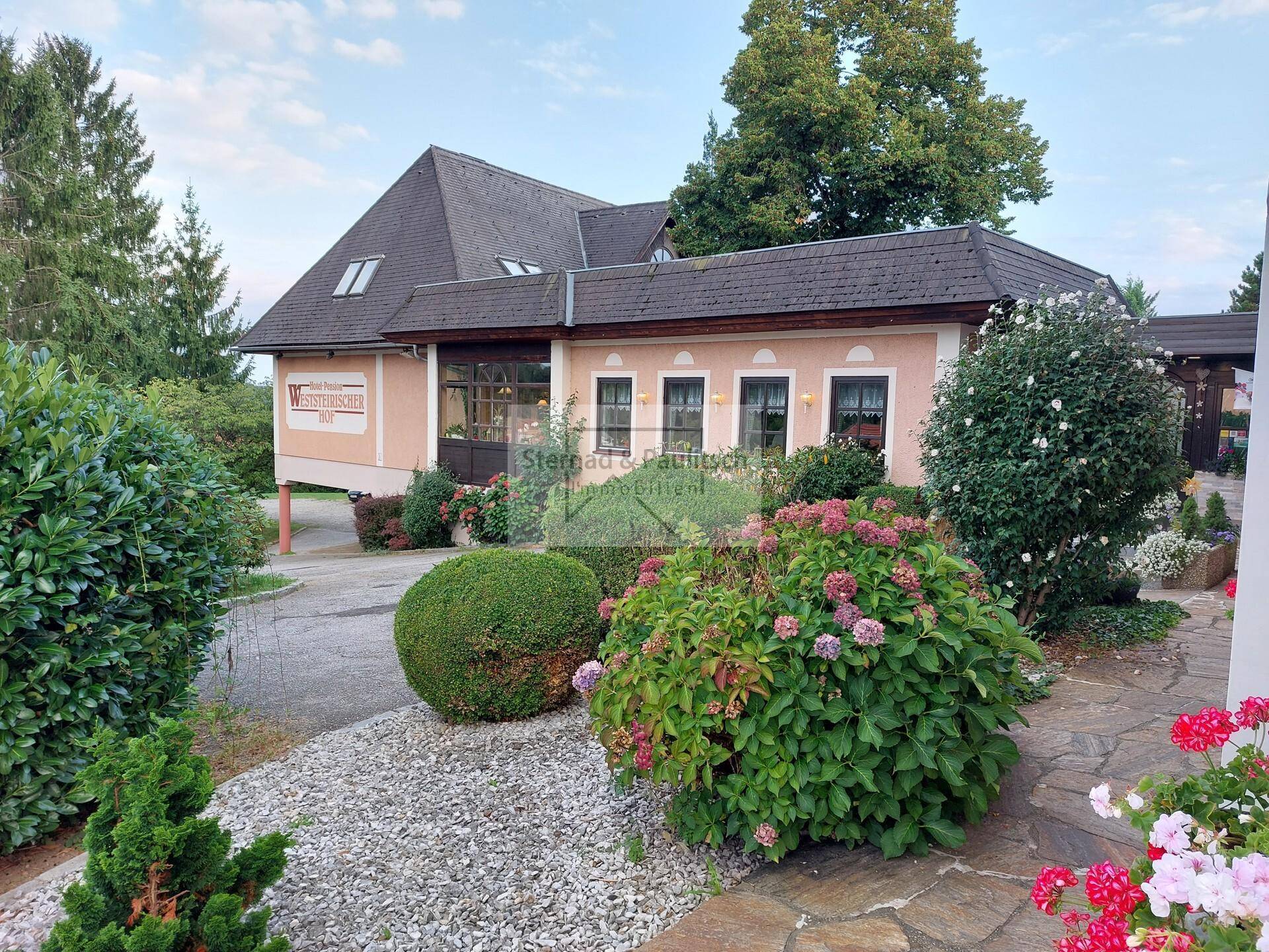Ansicht des Gebäudes mit gepflegtem Vorgarten und blühenden Sträuchern am Abend.