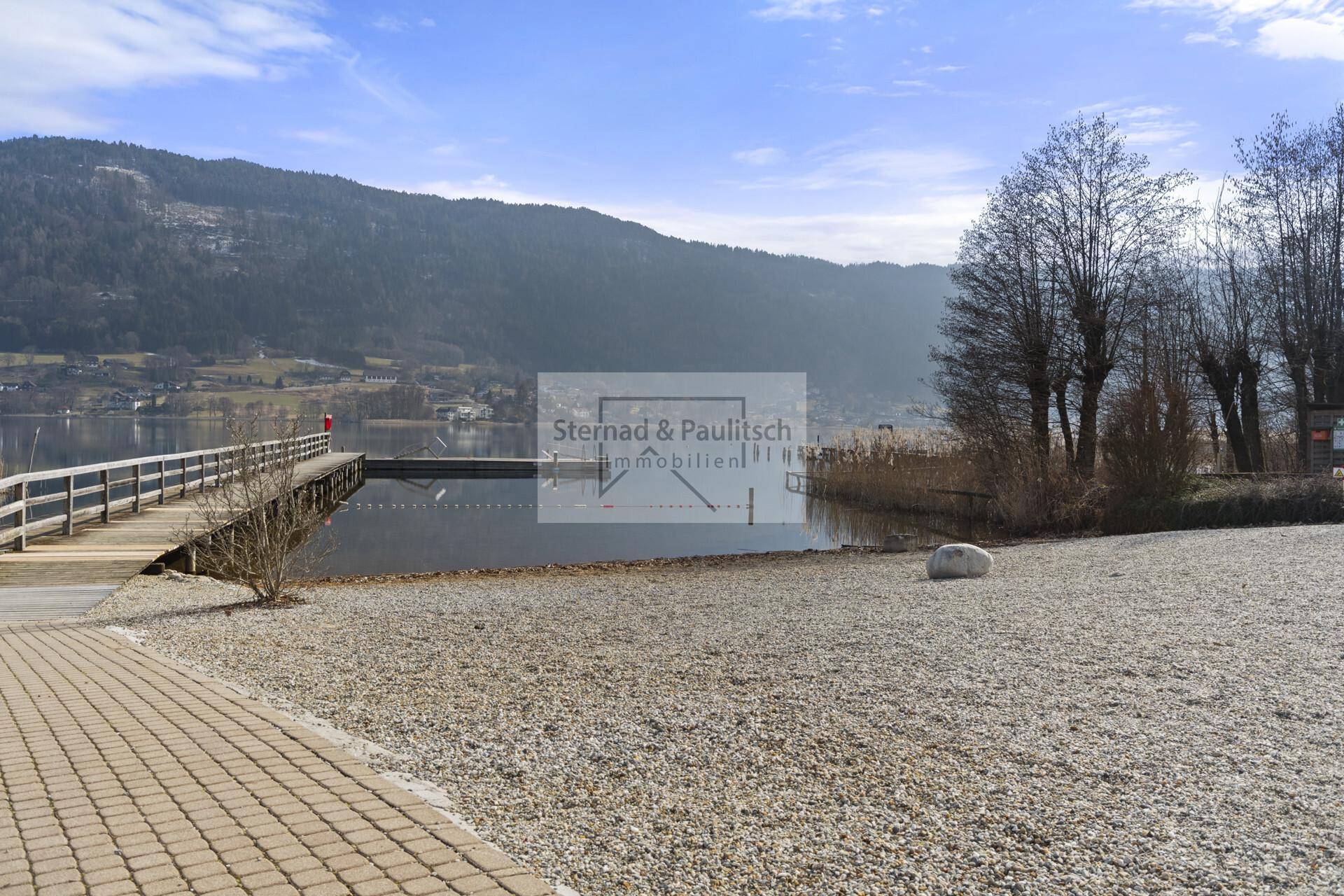 Kiesstrand am Seeufer mit Holzsteg und Blick auf die umliegende Berglandschaft unter blauem Himmel.