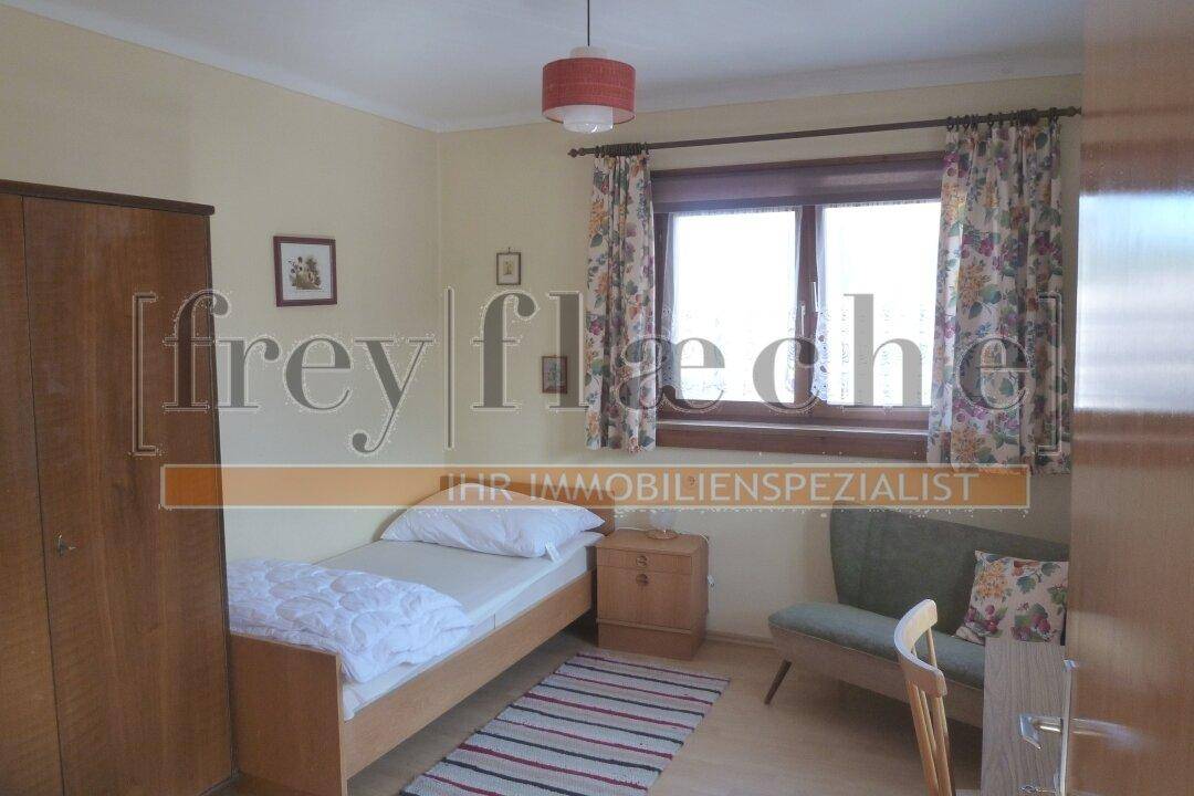 Einfaches Schlafzimmer mit Einzelbett, Holzschrank und Fenster mit floralen Vorhängen.