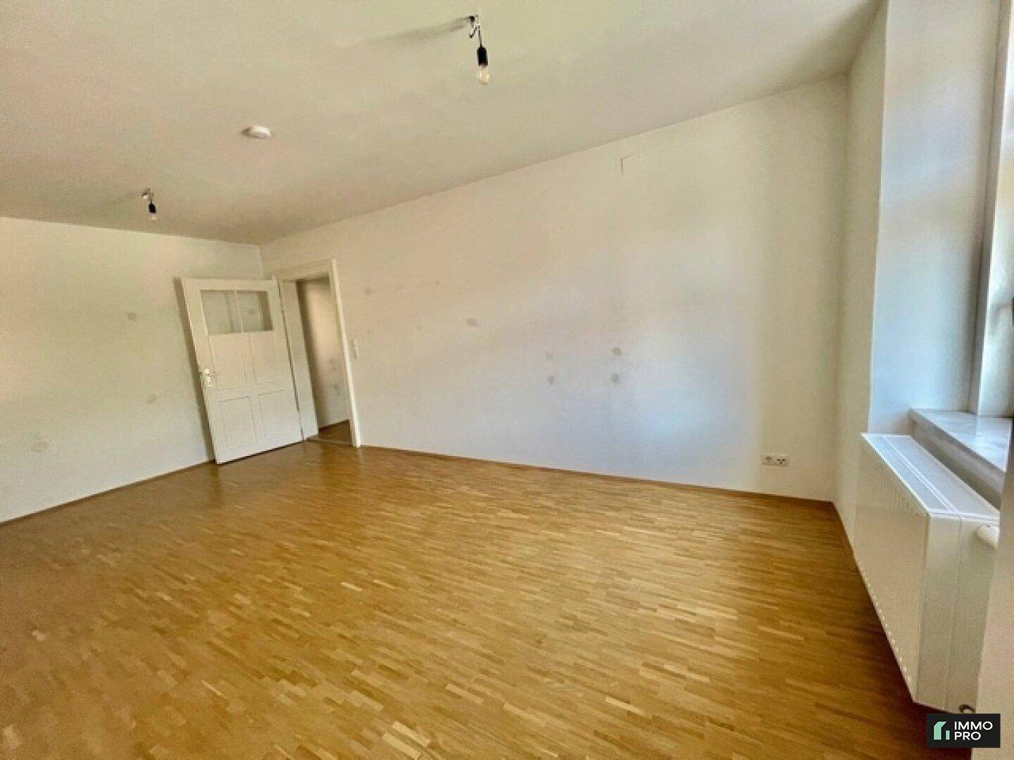 Geräumiger Raum mit Holzboden, weißen Wänden und natürlichem Lichteinfall durch ein Fenster.