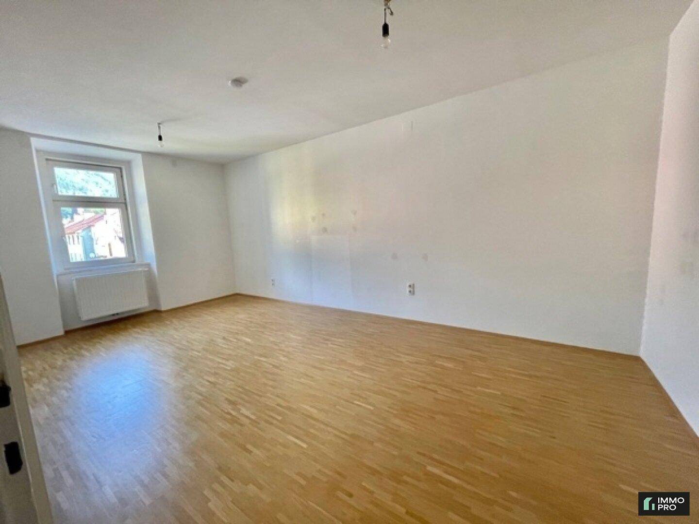 Leeres Wohnzimmer mit Holzboden, weißen Wänden und einem Fenster mit Heizkörper.
