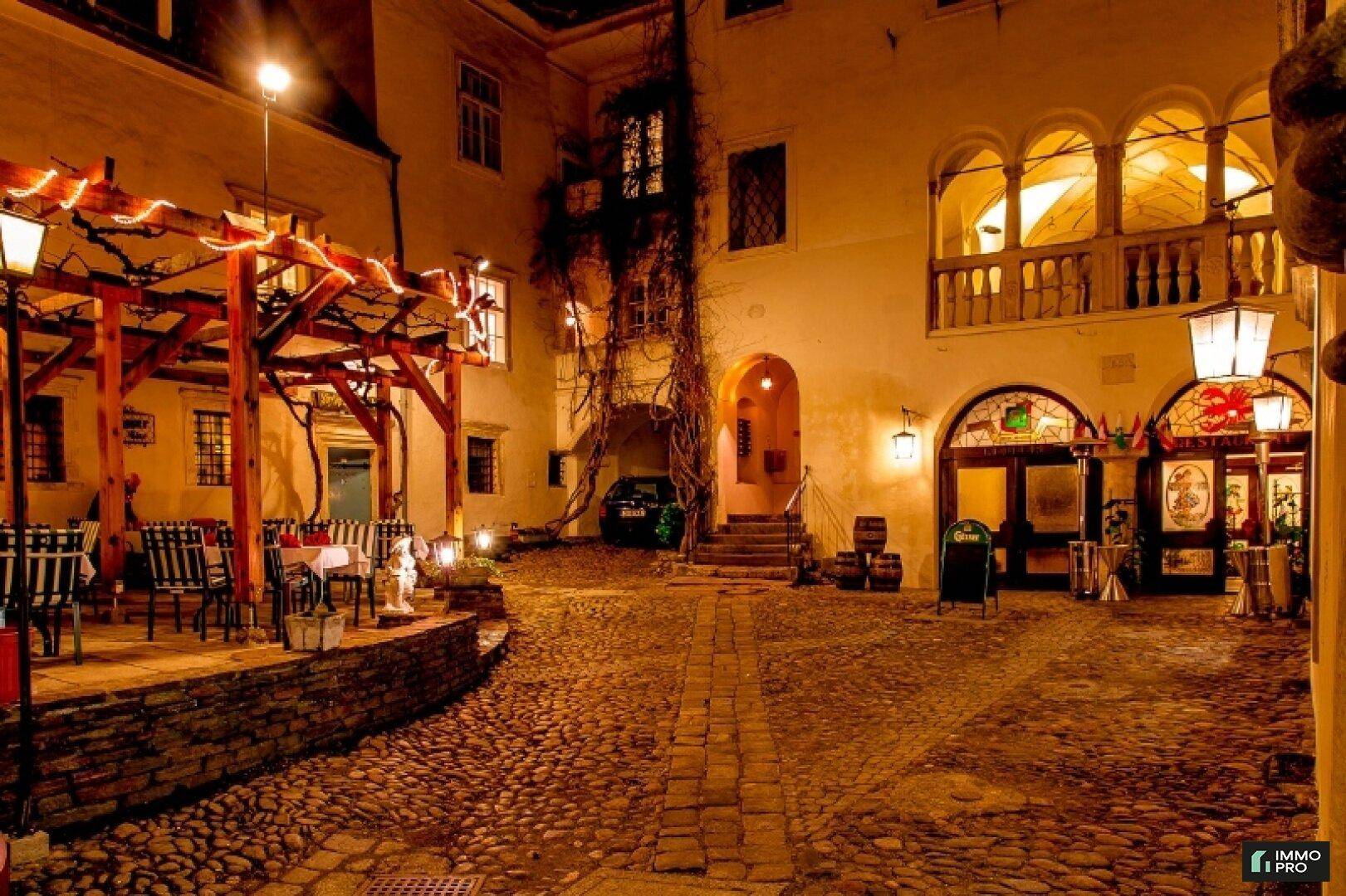 Romantischer Innenhof bei Nacht mit Kopfsteinpflaster, beleuchteter Pergola und gemütlichen Sitzgelegenheiten.