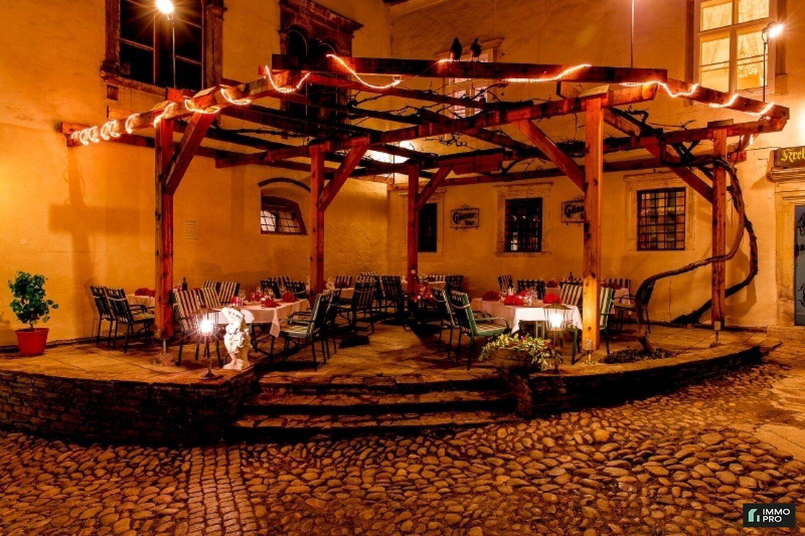 Beleuchteter Außenbereich mit Holzpergola und zahlreichen Tischen für Abendessen im Freien.