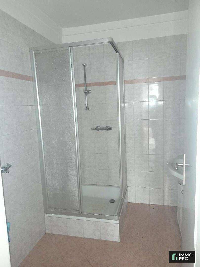 Badezimmer mit Duschkabine, hellen Fliesen und einem kleinen Waschbecken.