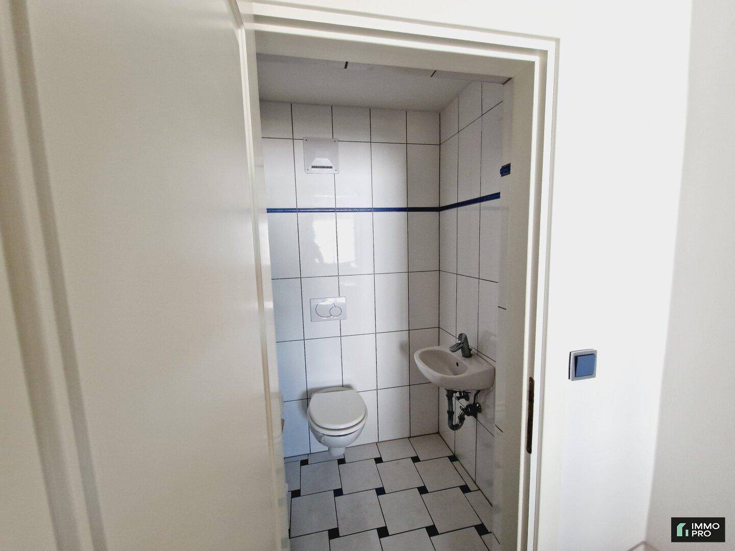 Kleines Gäste-WC mit weißen Fliesen, wandhängender Toilette und kleinem Waschbecken, sauber und funktional.