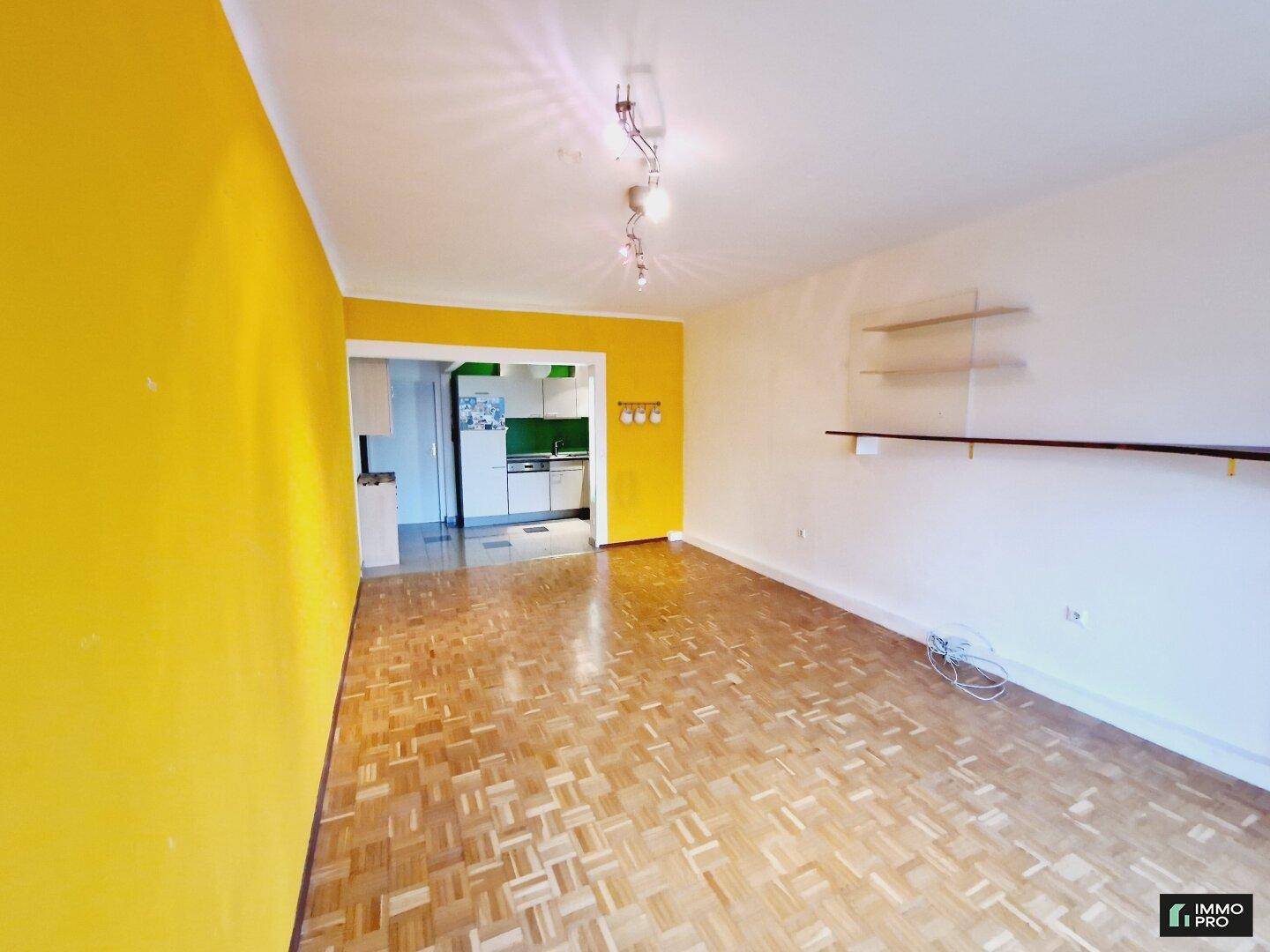 Geräumiges Wohnzimmer mit Parkettboden und einer auffälligen gelben Wand, Blick zur Küche.