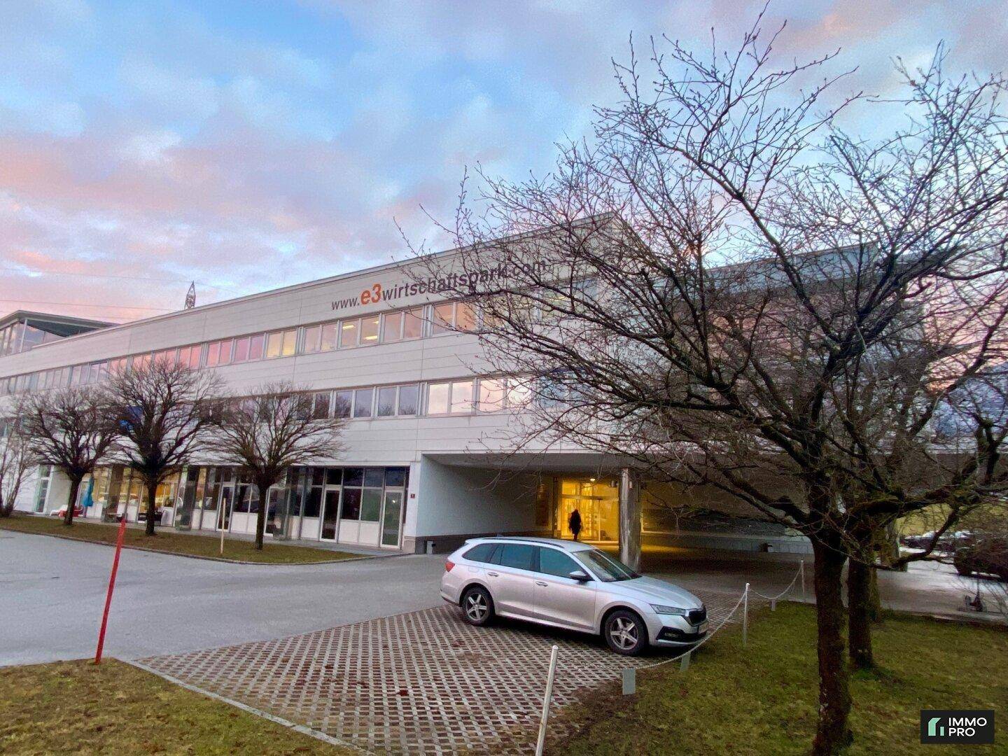 Außenansicht des modernen Bürogebäudes mit Parkplätzen und Bäumen im Vordergrund bei Sonnenuntergang.