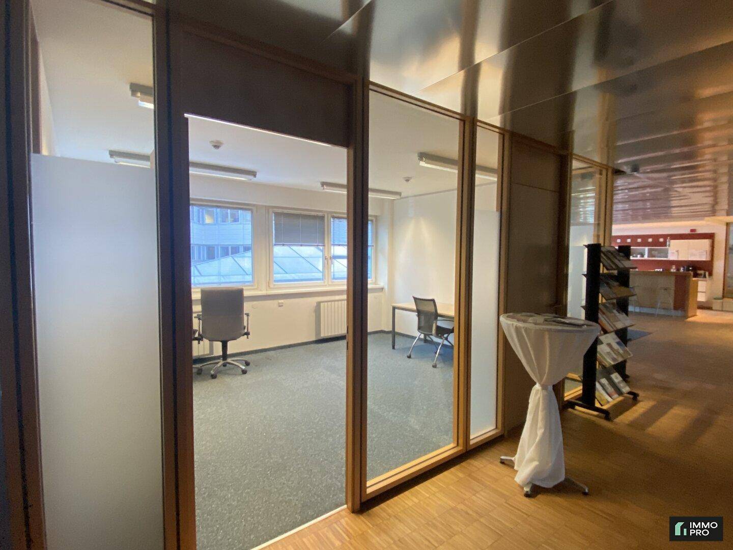 Helles Büro mit Teppichboden und großen Fenstern, sichtbar durch eine Glaswand vom Flur aus.