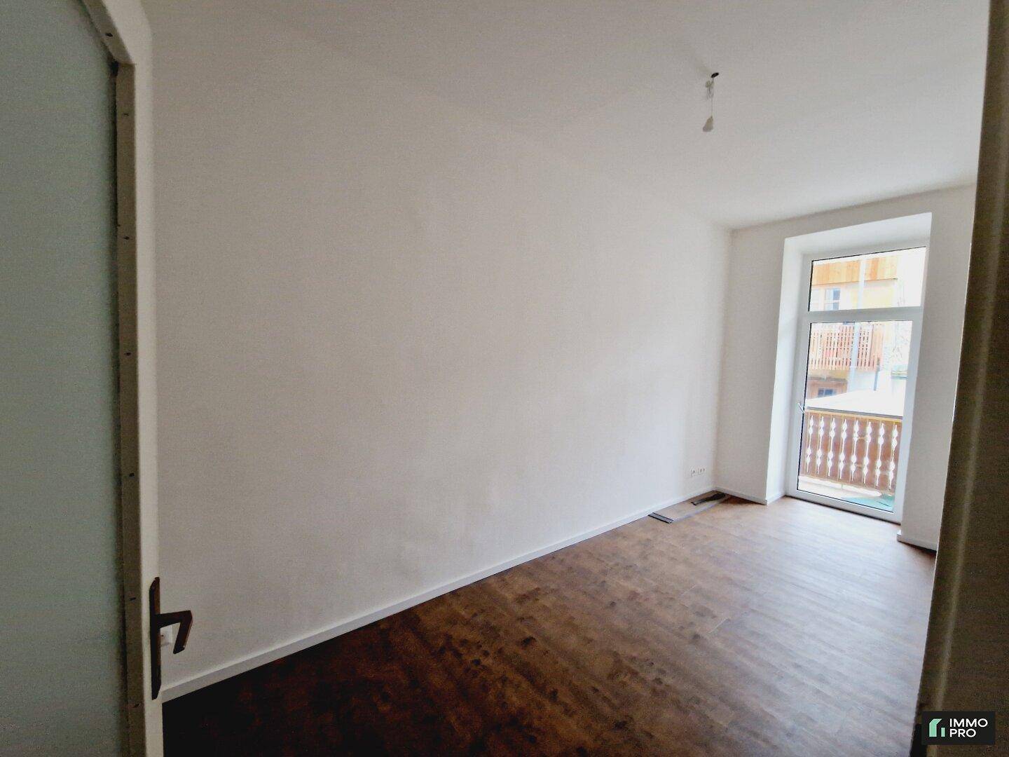 Helles Zimmer mit dunklem Holzboden und direktem Zugang zu einem Balkon durch eine große Glastür.
