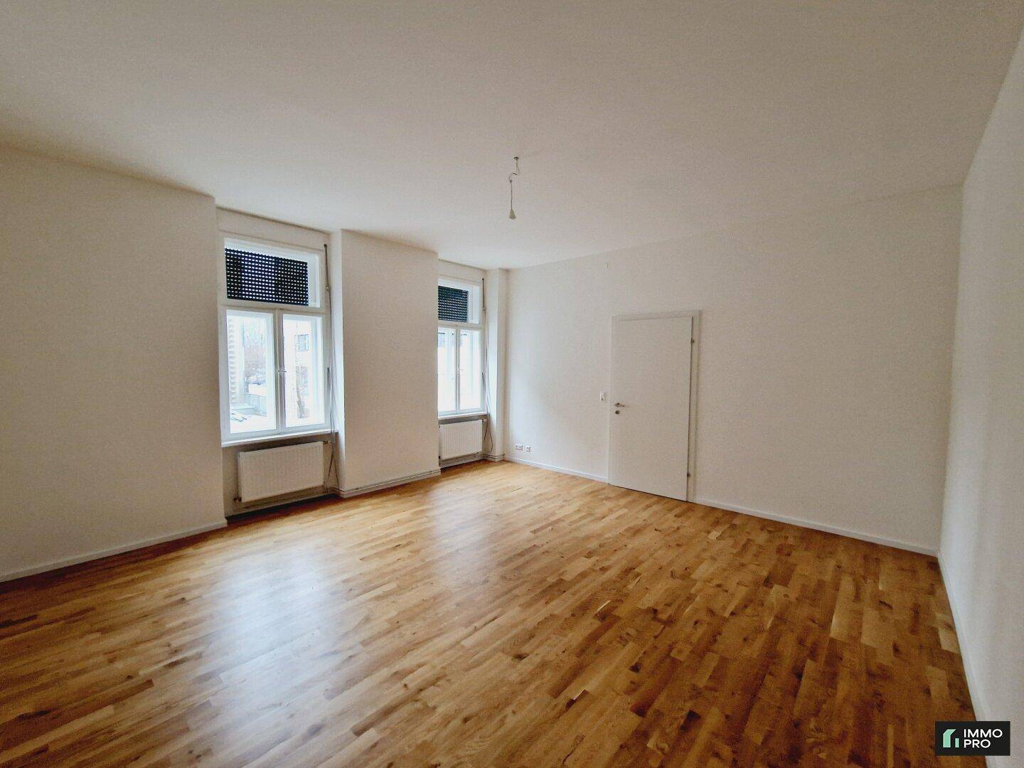 Geräumiges, leeres Wohnzimmer mit Holzboden, weißen Wänden und zwei Fenstern, die viel Licht hereinlassen.