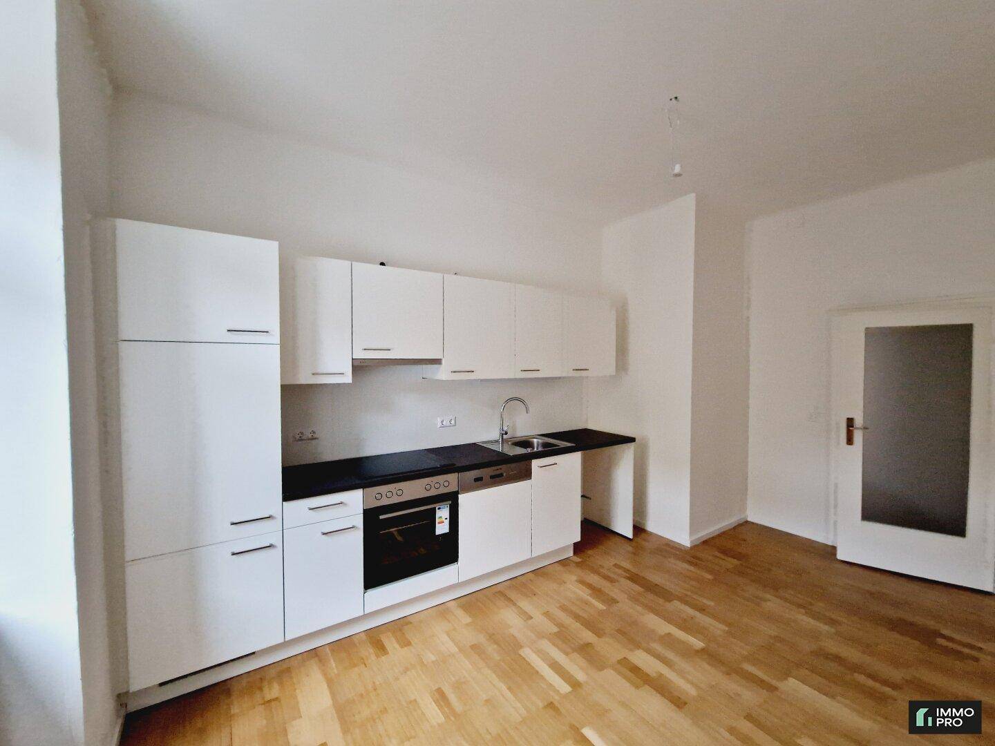 Funktionale Küchenzeile mit weißen Fronten, Spüle und Backofen, angrenzend an einen Raum mit Holzboden.