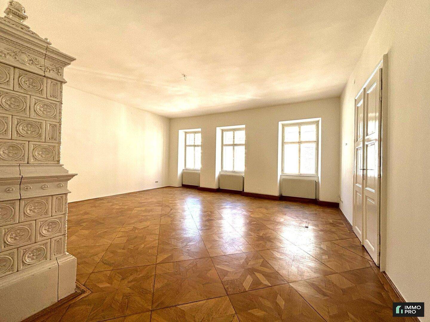 Großzügiger Raum mit drei hohen Fenstern und einem eleganten Kachelofen, der viel Tageslicht hereinlässt.