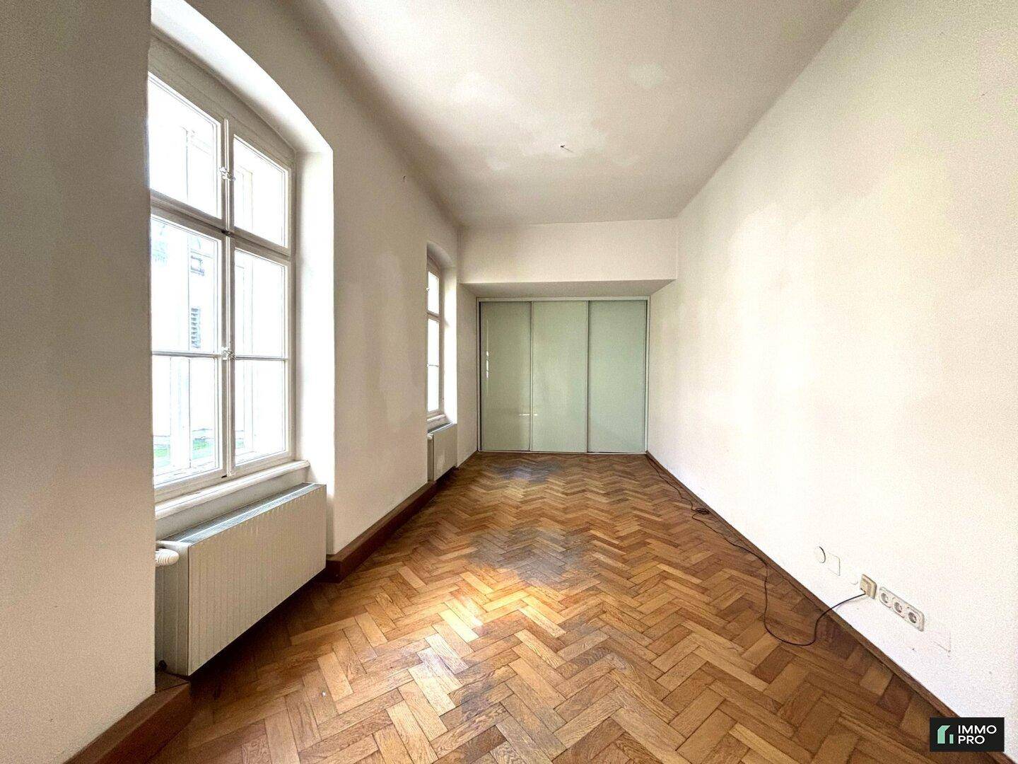 Langes Zimmer mit Fischgrätparkett, großem Fenster und einem praktischen Einbauschrank mit Schiebetüren.