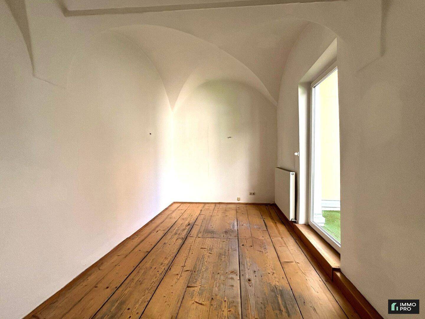 Schmaler Raum mit rustikalem Holzboden, einem großen Fenster und einer einzigartigen Gewölbedecke.