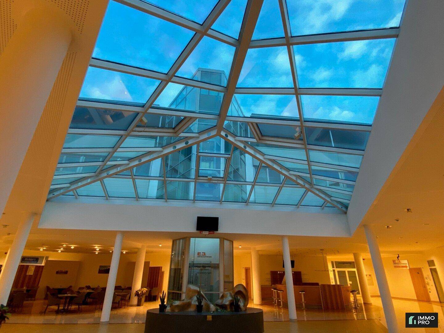 Blick nach oben auf das beeindruckende Glasdach und den transparenten Aufzug in der modernen Lobby.