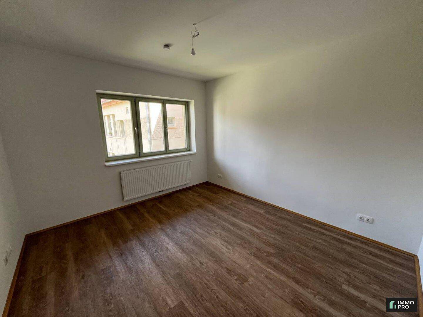 Leeres Schlafzimmer mit Laminatboden, weißen Wänden und einem großen Fenster mit grünem Rahmen.