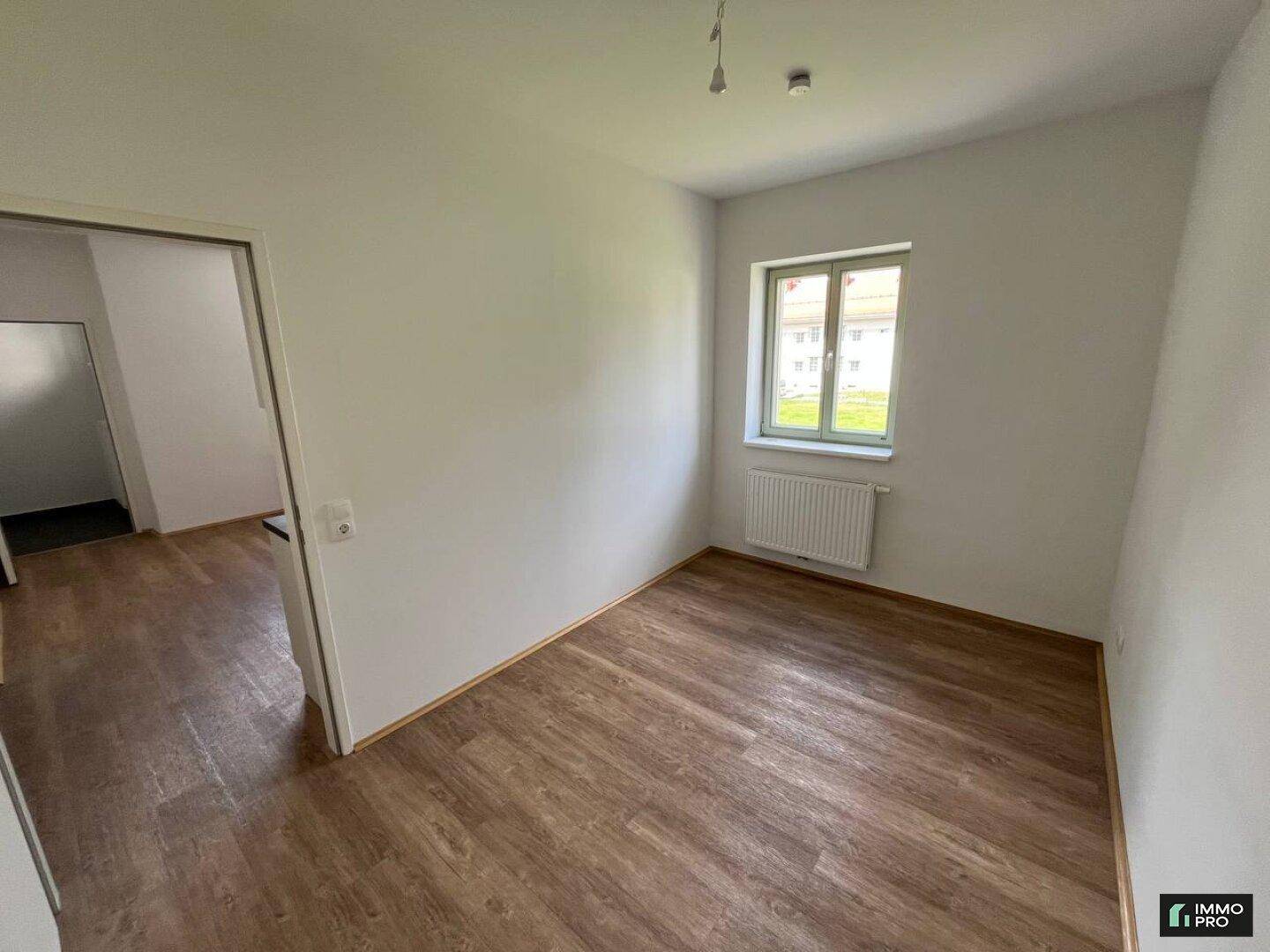 Helles Zimmer mit Laminatboden, weißen Wänden und einem Fenster, das einen Blick nach draußen bietet.