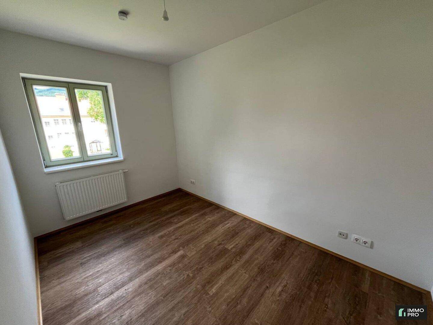 Geräumiges Zimmer mit Laminatboden, weißen Wänden und einem Fenster, das viel Tageslicht hereinlässt.