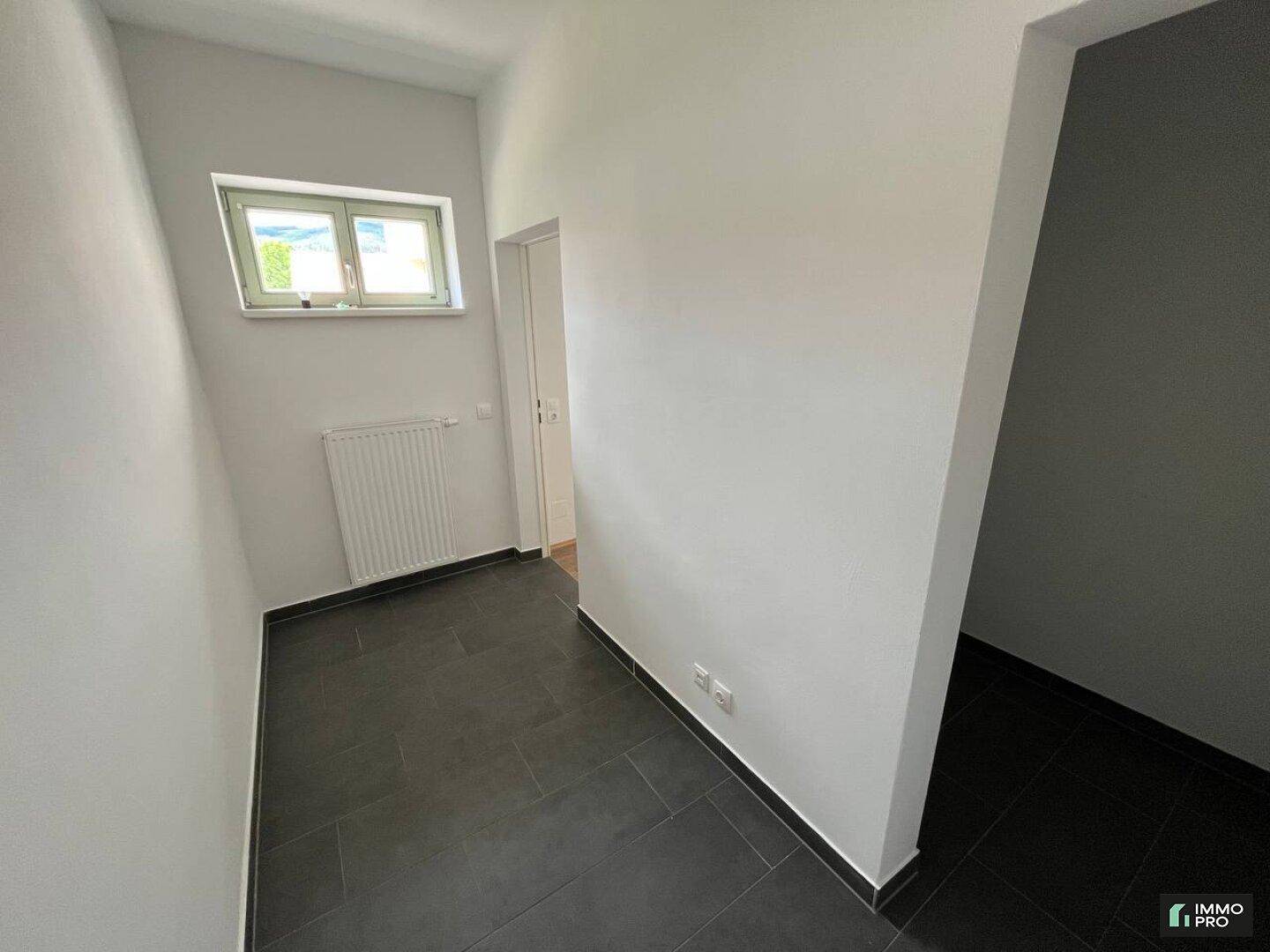 Heller Flur mit dunklen Fliesen, weißen Wänden und einem Fenster, das Tageslicht spendet.