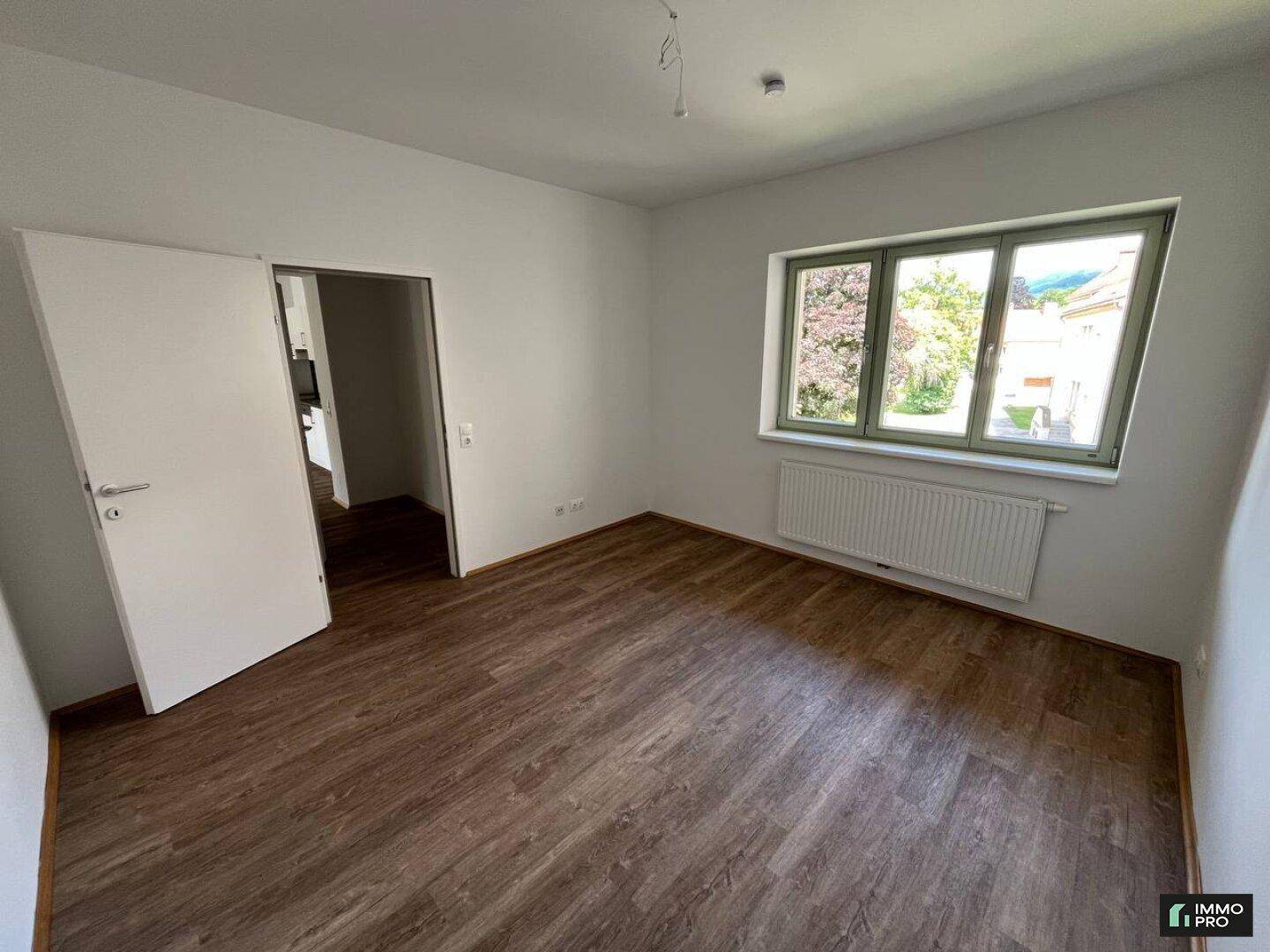 Freundliches Zimmer mit Laminatboden, weißen Wänden und einem großen Fenster mit Blick ins Grüne.