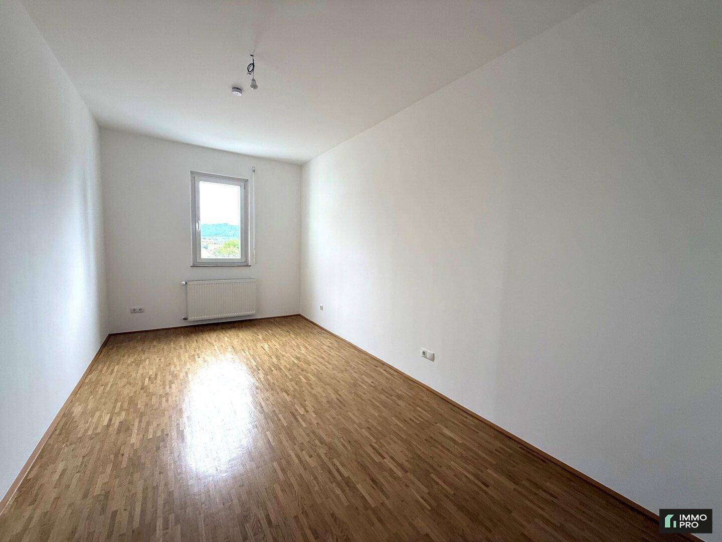 Leeres Zimmer mit Parkettboden und einem Fenster, das viel Tageslicht hereinlässt.