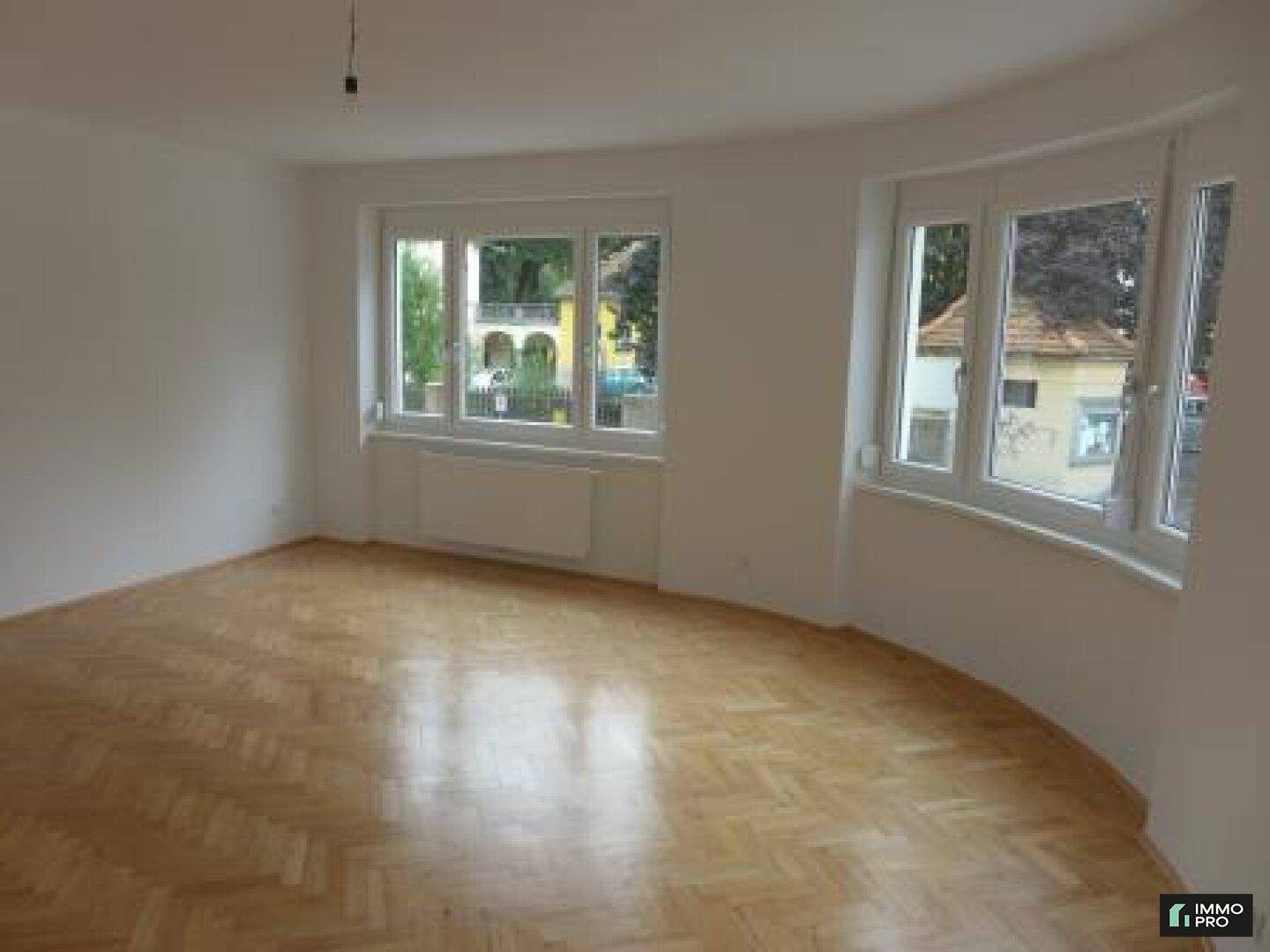 Lichtdurchfluteter Raum mit Fischgrätparkett und weiten Fenstern, ideal für Wohnzwecke.
