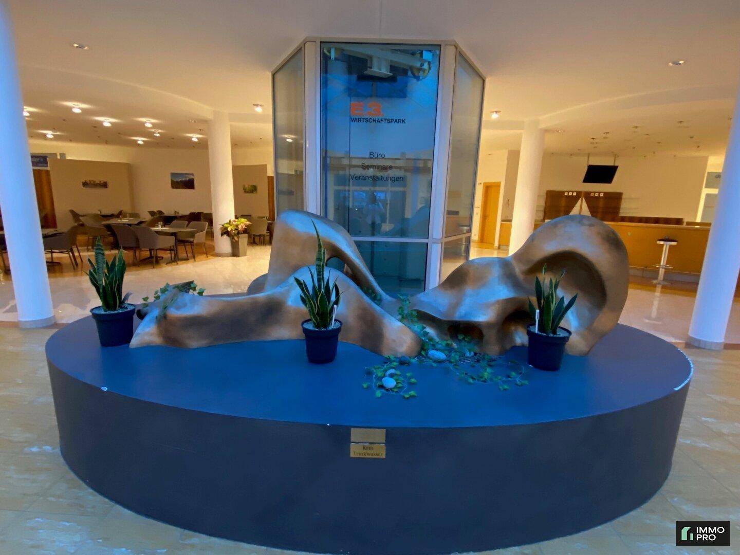Empfangsbereich mit moderner Skulptur, Pflanzen und Blick auf den angrenzenden Essbereich.