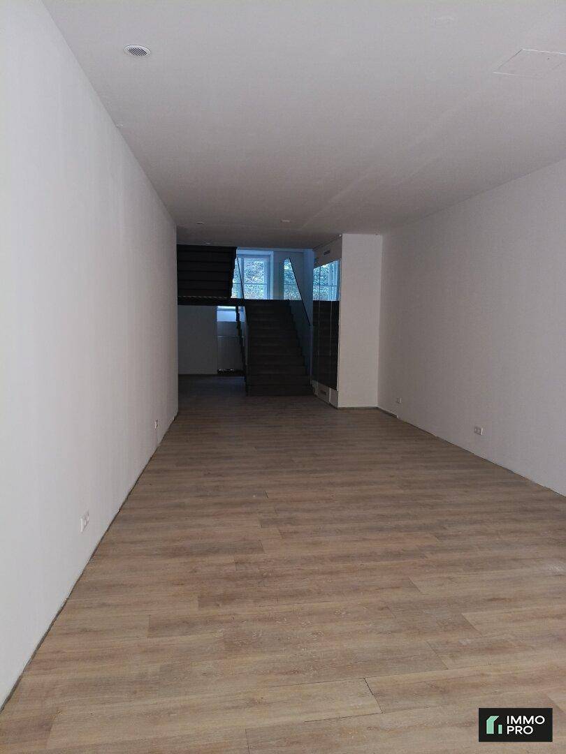 Heller, leerer Raum mit hellem Laminatboden und Blick auf eine moderne Treppe im Hintergrund.