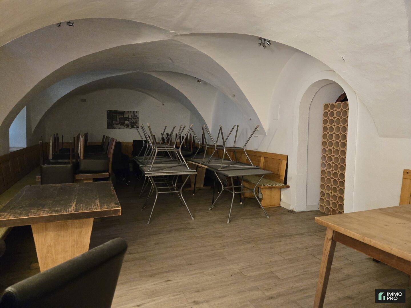 Gastraum mit gewölbter Decke, Holzbänken und Tischen, sowie gestapelten Stühlen, bereit zur Nutzung.