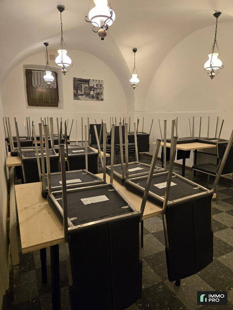 Gastraum mit kariertem Fliesenboden, Holztischen und gestapelten Stühlen unter historischen Deckenleuchten.