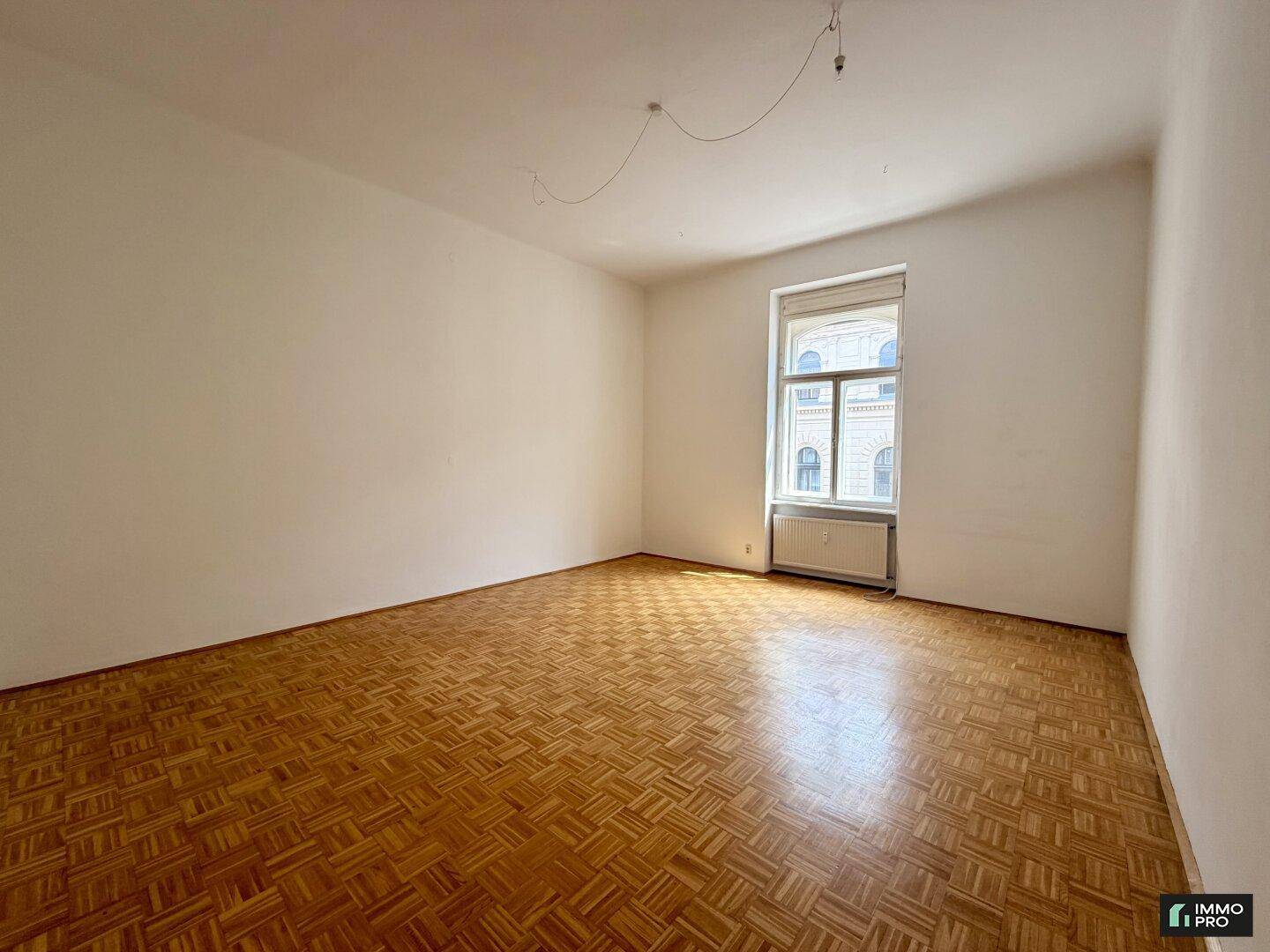 Helles Zimmer mit Fischgrätparkettboden und einem Fenster, das Tageslicht hereinlässt.