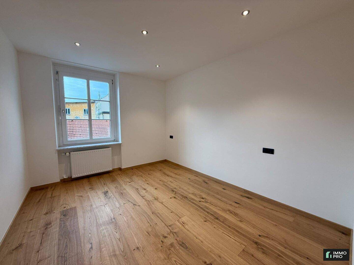 Leeres Schlafzimmer mit Holzboden, weißen Wänden und einem großen Fenster für viel Tageslicht.