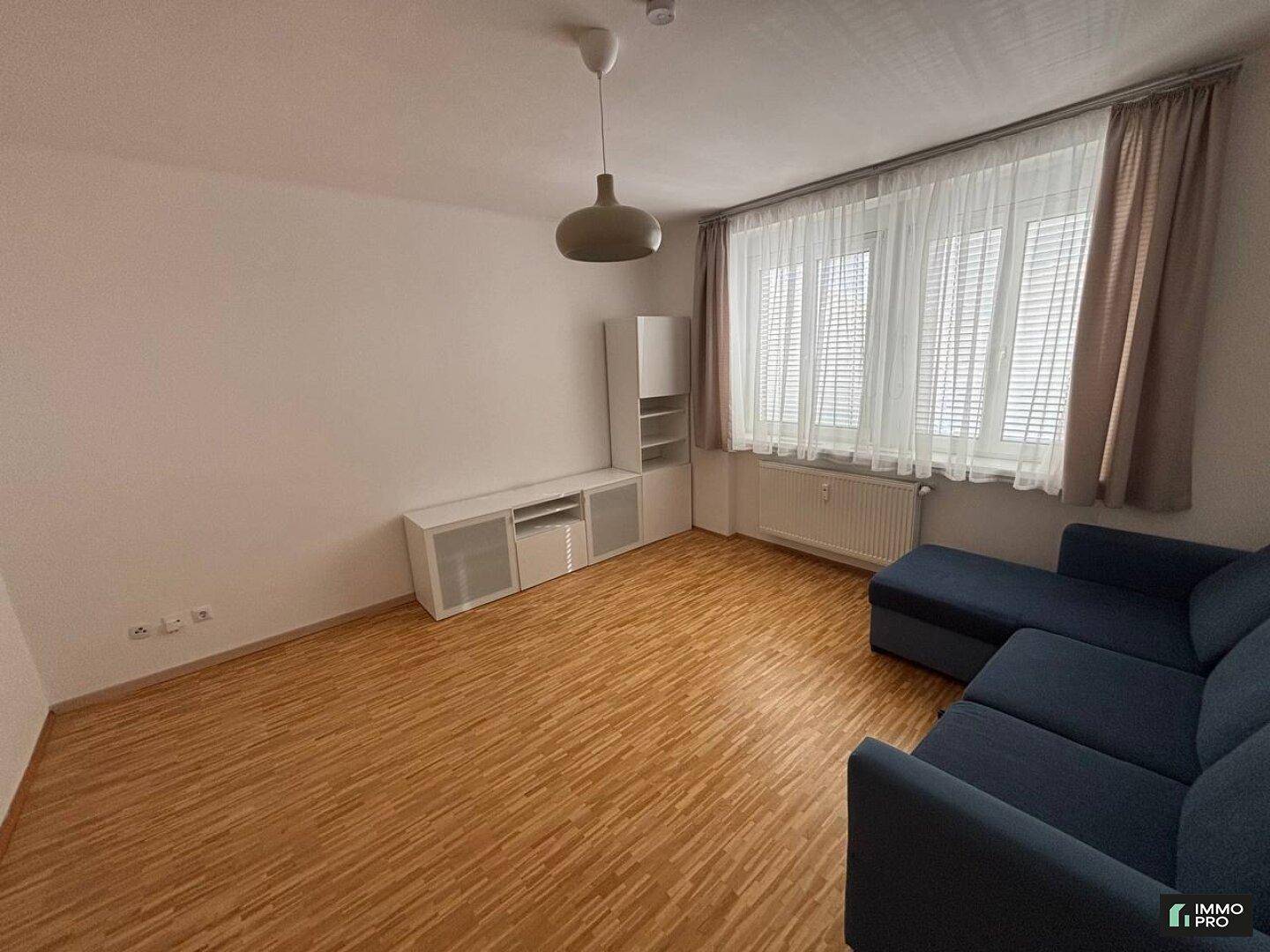 Geräumiges Wohnzimmer mit Holzboden, blauer Eckcouch und großem Fenster.