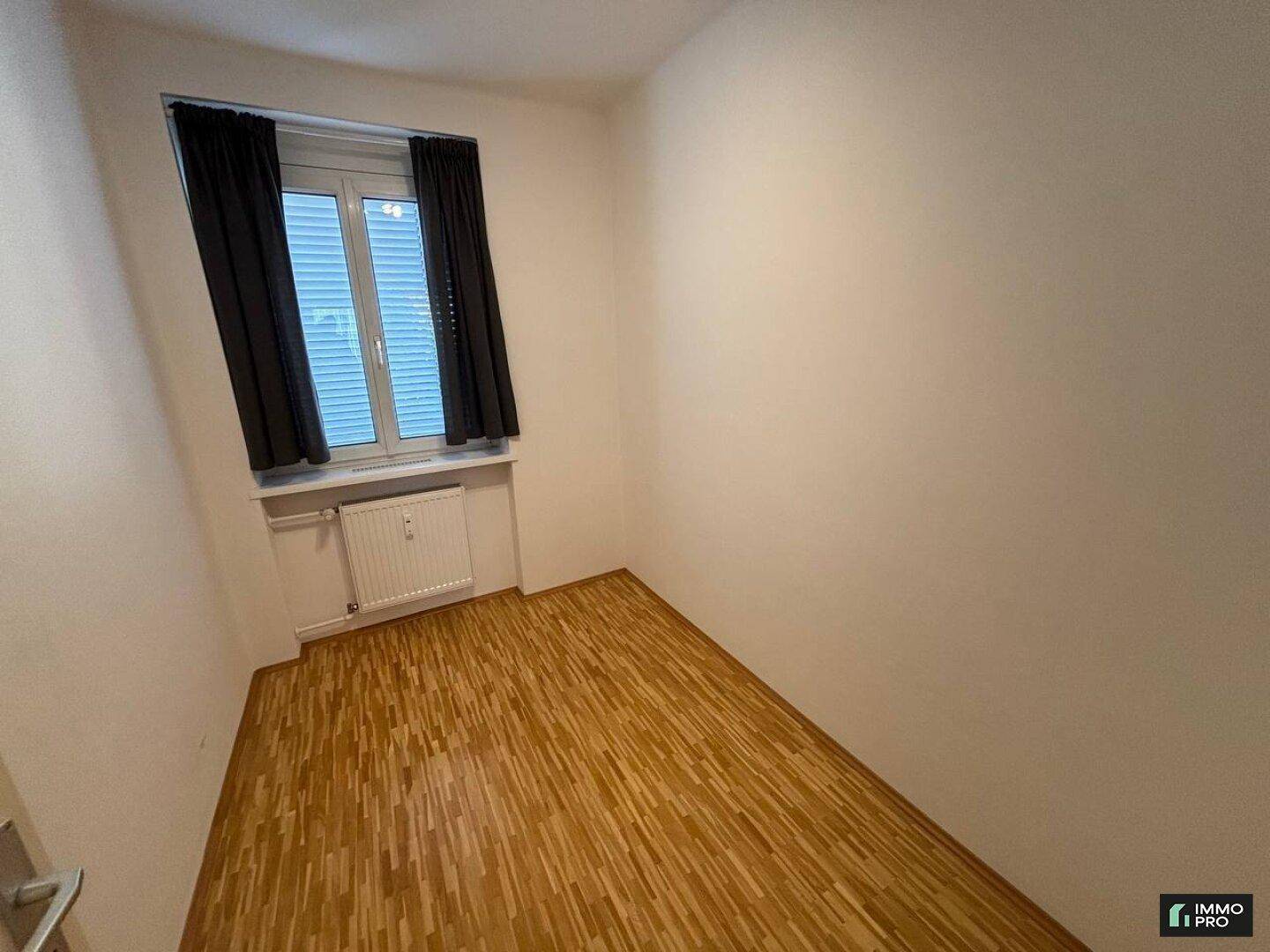 Kleines Schlafzimmer mit Parkettboden und Fenster mit dunklen Vorhängen.