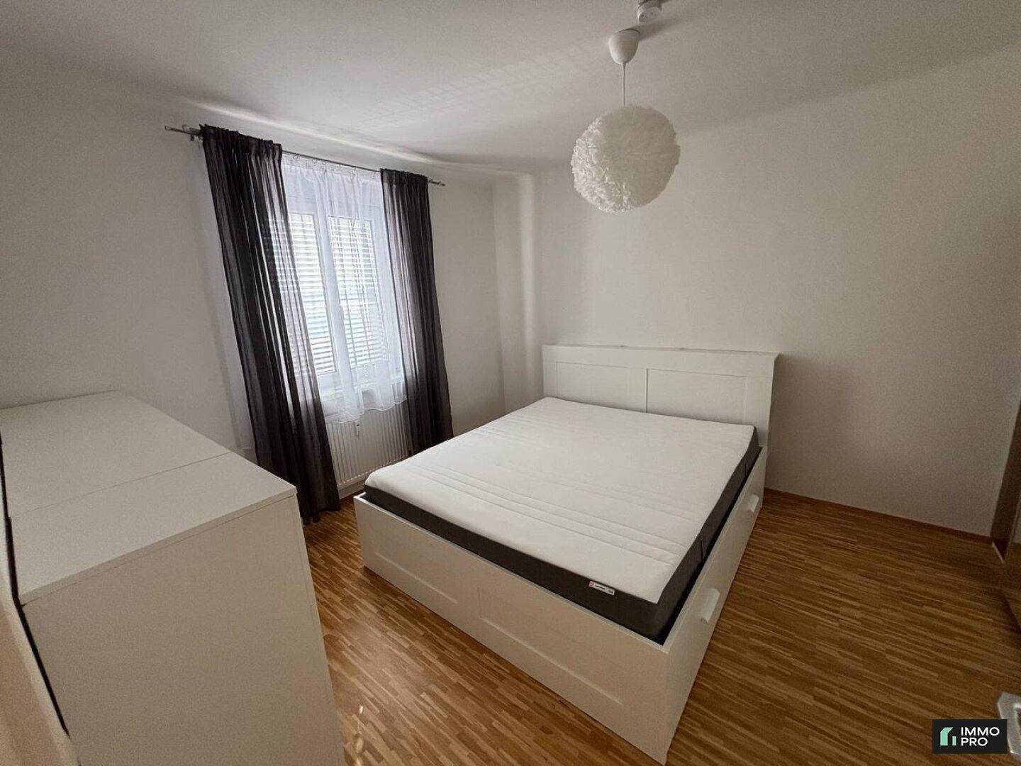 Schlafzimmer mit Doppelbett, weißer Kommode und Fenster mit dunklen Vorhängen.