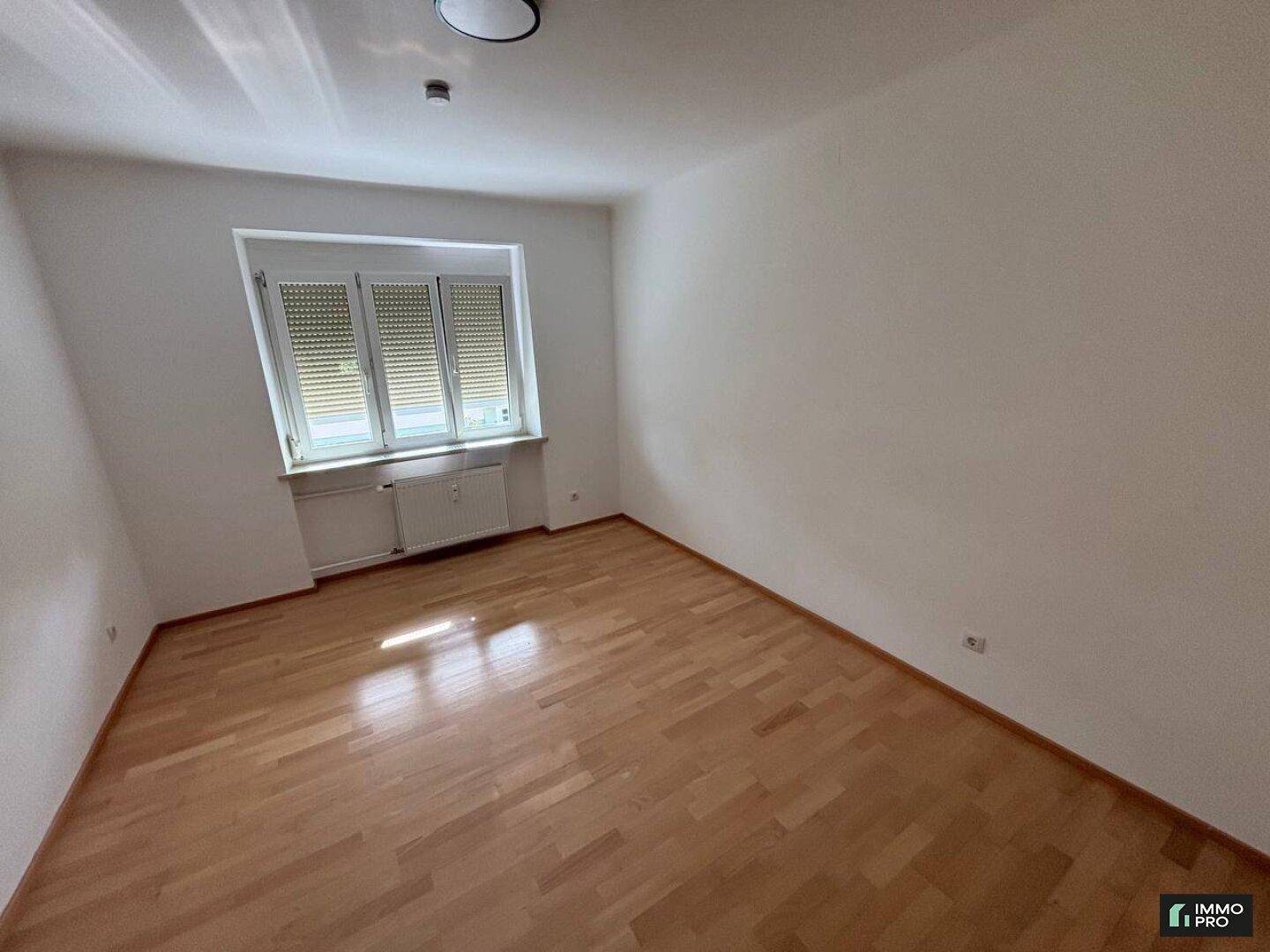 Leeres Schlafzimmer mit Holzfußboden, einem Fenster mit Rollläden und einem Heizkörper.