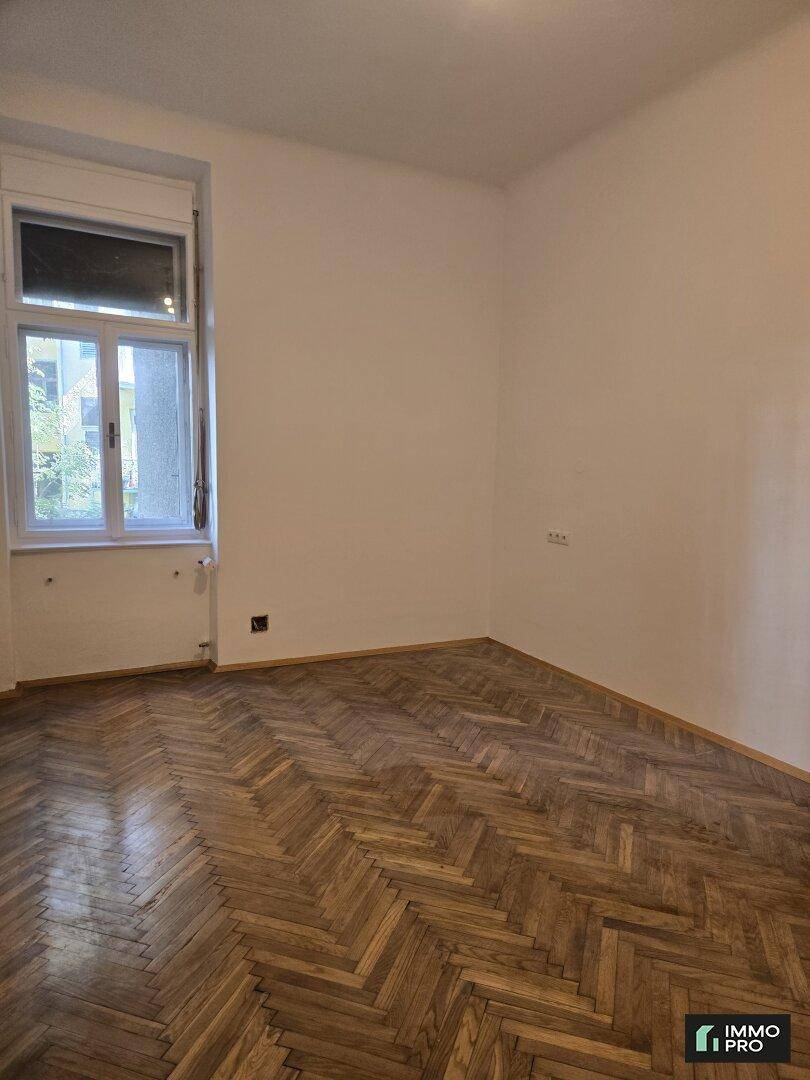 Leeres Zimmer mit schönem Fischgrätparkett und einem großen Fenster, das den Raum hell erscheinen lässt.