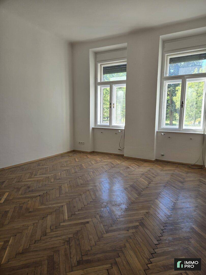 Helles, leeres Zimmer mit klassischem Fischgrätparkett und zwei großen Fenstern, die viel Licht hereinlassen.
