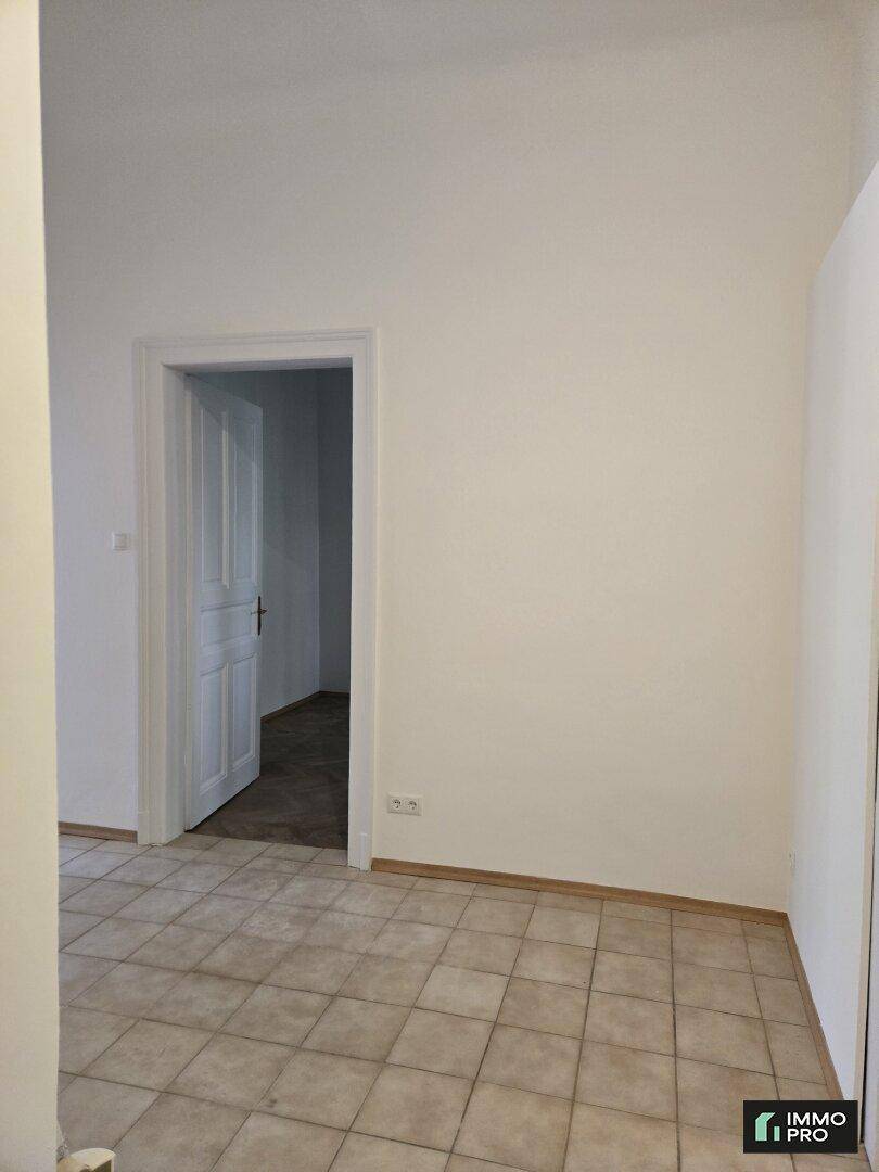 Leerer Raum mit hellen Fliesen und einer offenen Tür, die zu einem weiteren Bereich der Wohnung führt.