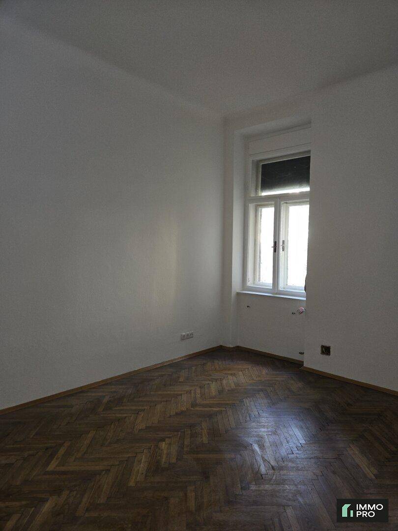 Geräumiges Zimmer mit Fischgrätparkett und einem großen Fenster, das den Raum mit Tageslicht erfüllt.