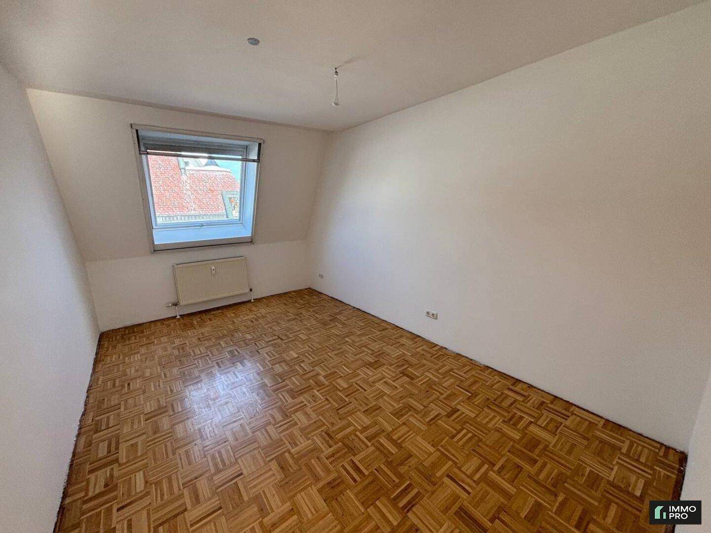 Leeres Zimmer mit Parkettboden, weißen Wänden und einem Dachfenster für natürliches Licht.